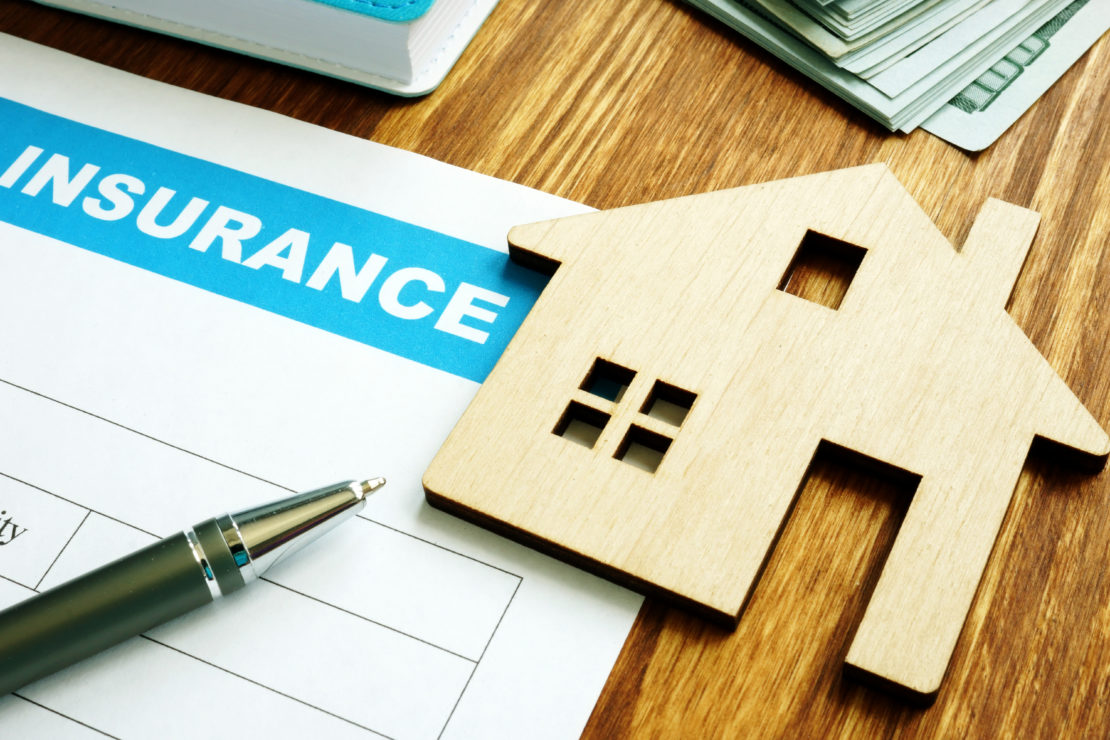 House insurance form for homeowner. A flat, wooden model of a home is laying on top of the form. A pen also sits on top of the home insurance form.
