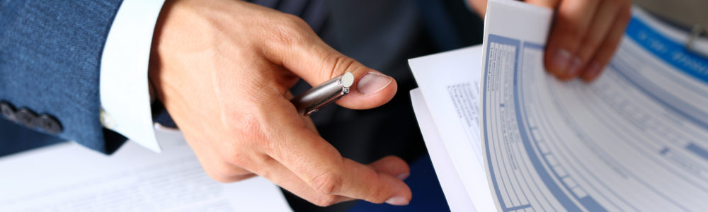 close up of a man in a blue suit holding a silver pen in one hand and a stack of insurance claims paper in the other hand