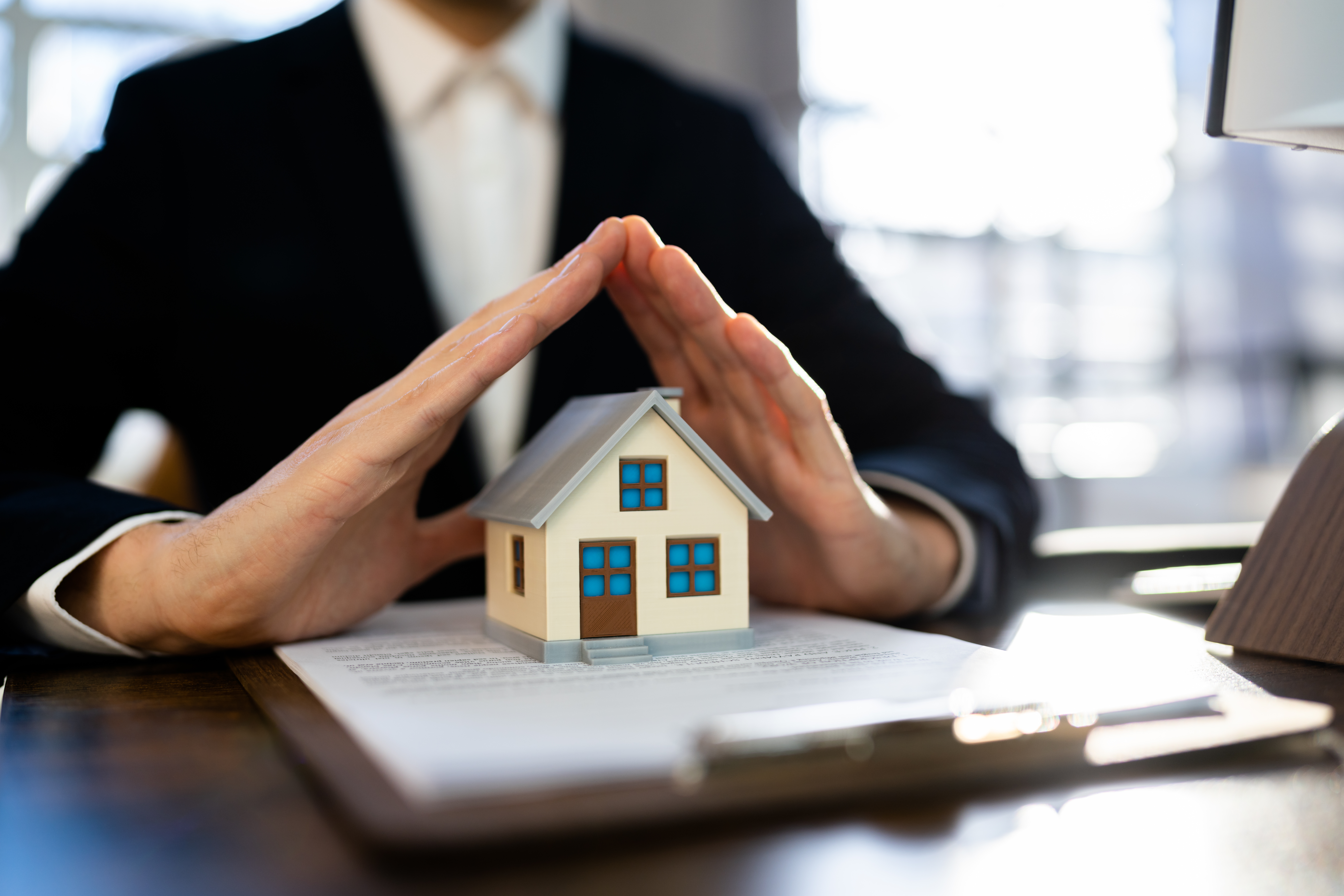 a photo of a suited insurance agent (blurred in the background) hovering his hands over a tiny house and clipboard. 