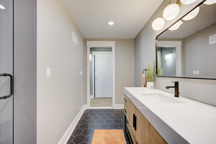 Modern bathroom featuring a custom white oak vanity, integrated white solid surface countertop, matte black faucet, black hexagon floor tile, and frameless glass shower.