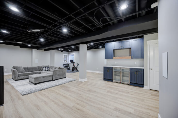 Luxury finished basement featuring a custom navy wet bar with beverage refrigerators, quartz countertops, luxury vinyl plank flooring, black painted exposed ceiling, and open family entertainment area.