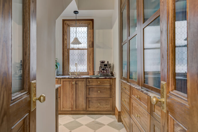 Elegant butler's pantry featuring custom stained Dura Supreme inset cabinetry, marble countertops, leaded glass window, unlacquered brass sink and faucet, and checkered marble tile flooring.