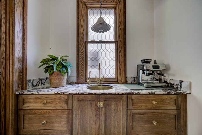 Luxury butler's pantry featuring stained Dura Supreme inset cabinetry, marble countertops, unlacquered brass sink and faucet, fluted glass cabinets, and green and white marble tile flooring.