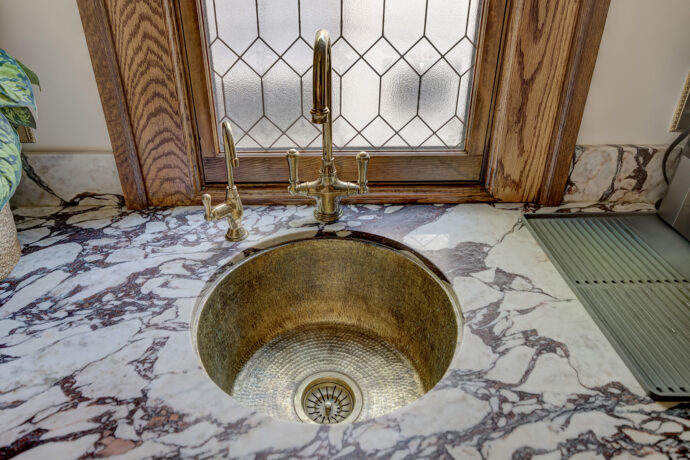 High-end Birmingham butler's pantry featuring Dura Supreme inset cabinetry, natural marble countertops, green and white checkerboard marble flooring, unlacquered brass sink and faucet, and custom fluted glass storage.