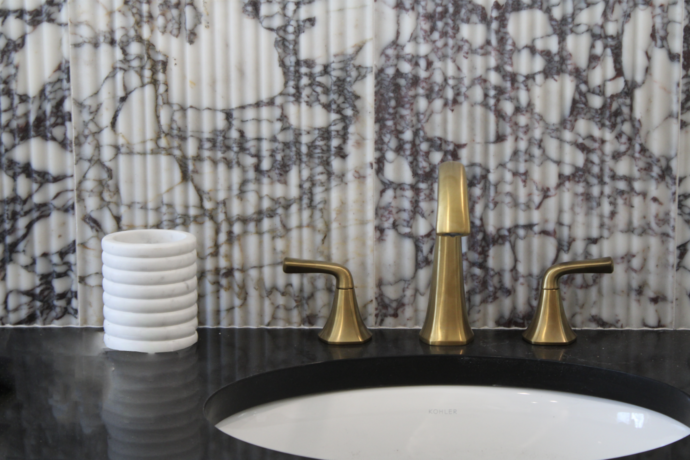 Close-up of a luxury bathroom vanity featuring a sculptural brushed brass faucet, black quartz countertop, undermount sink, and dramatic fluted marble backsplash.
