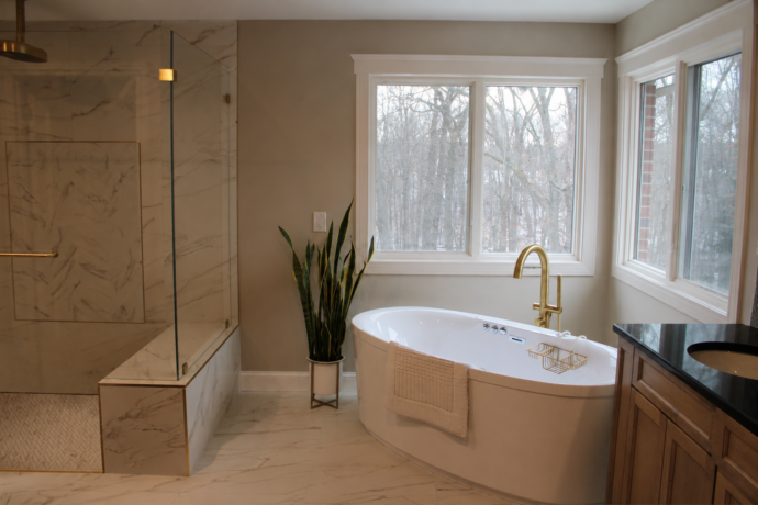 Luxury primary bathroom featuring a freestanding soaking tub, curbless marble shower, frameless glass enclosure, brass fixtures, custom window casing, and porcelain tile flooring.