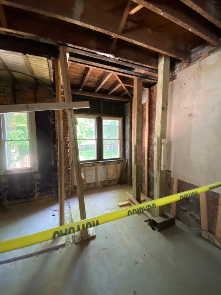 Interior demolition in a 1906 Detroit Tudor showing exposed brick walls, original ceiling joists, temporary wood shoring posts and a new steel beam overhead.