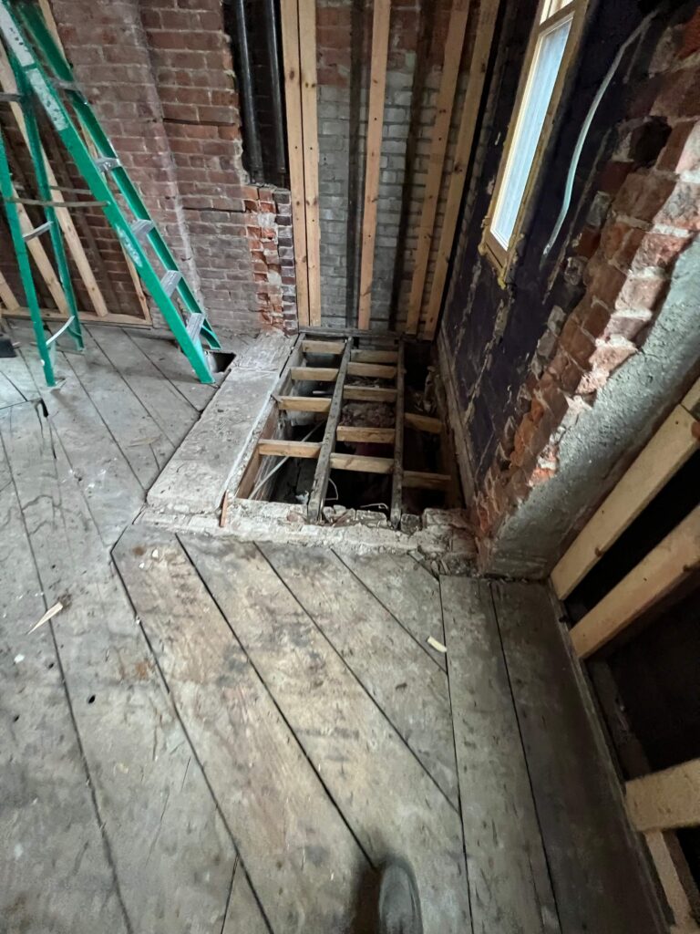 Original floor joists and wide plank subfloor exposed against a solid brick wall during renovation of a 1906 Detroit Tudor.