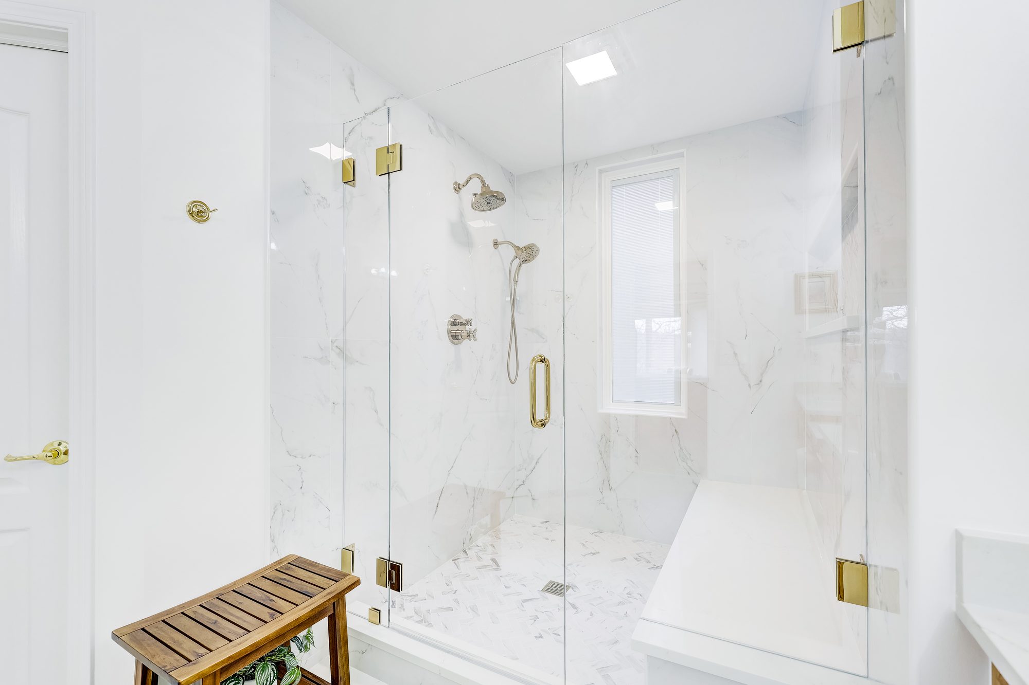 Frameless glass shower with polished brass hinges and a teak stool in a Birmingham MI bathroom remodel