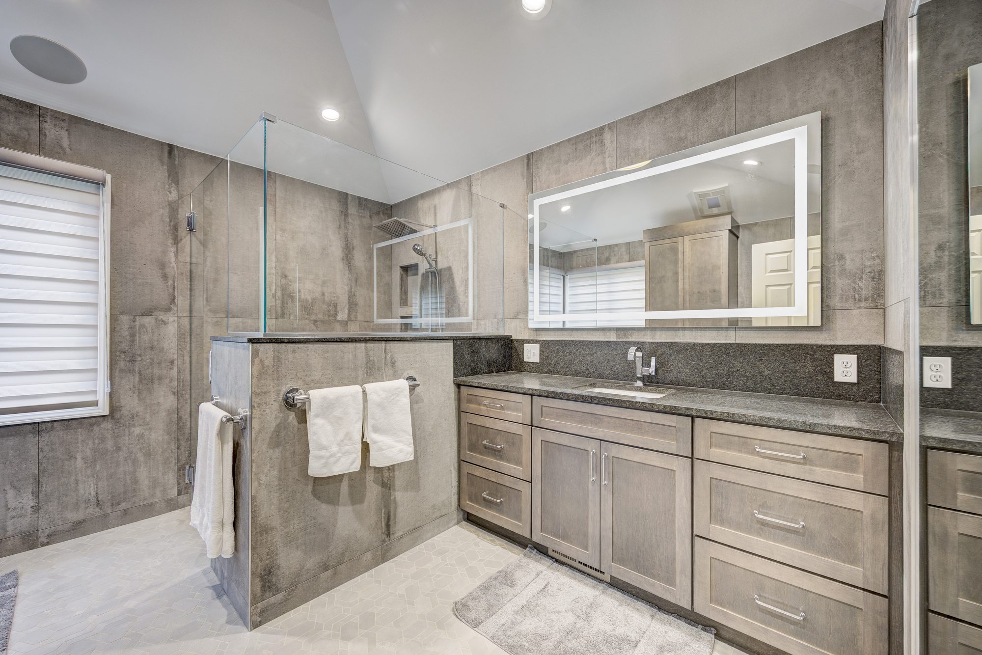 Remodeled bathroom in Troy MI with a curbless walk in shower, concrete look porcelain walls and a backlit LED mirror