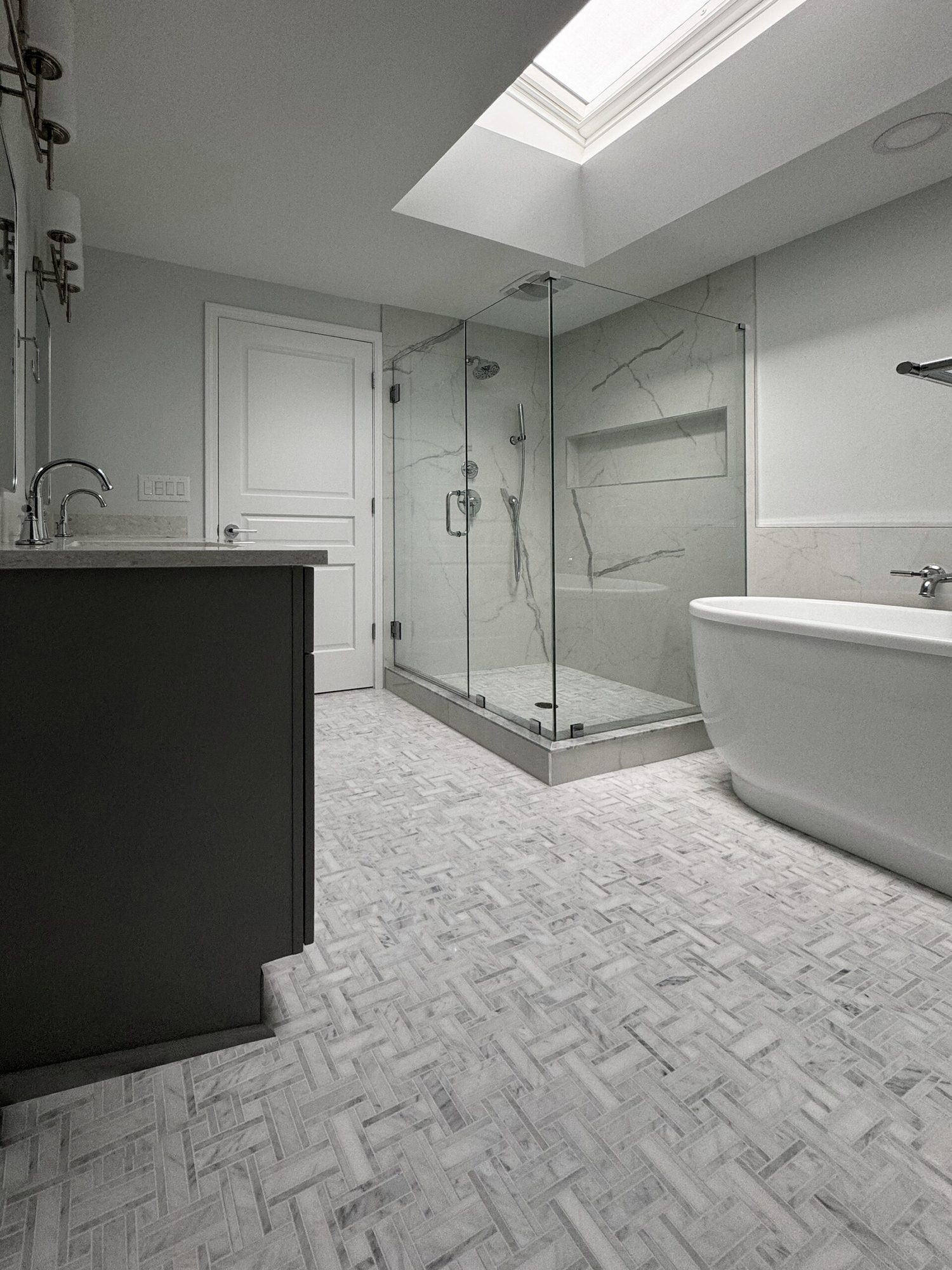 Remodeled bathroom in Troy MI with a frameless glass shower under a skylight, freestanding tub and marble herringbone floor