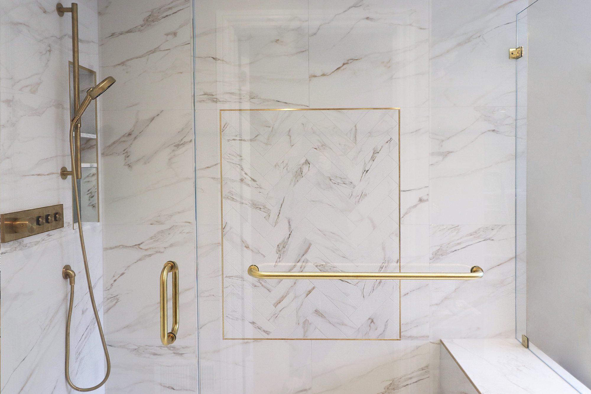 Shower wall with a herringbone tile inset framed in brass trim beside a brass hand shower in a Rochester Hills MI bathroom