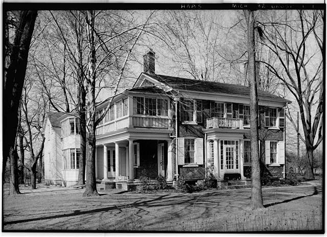Black and white photo of a two story brick Colonial with shutters, covered front porches and a clapboard wing, framed by bare trees.
